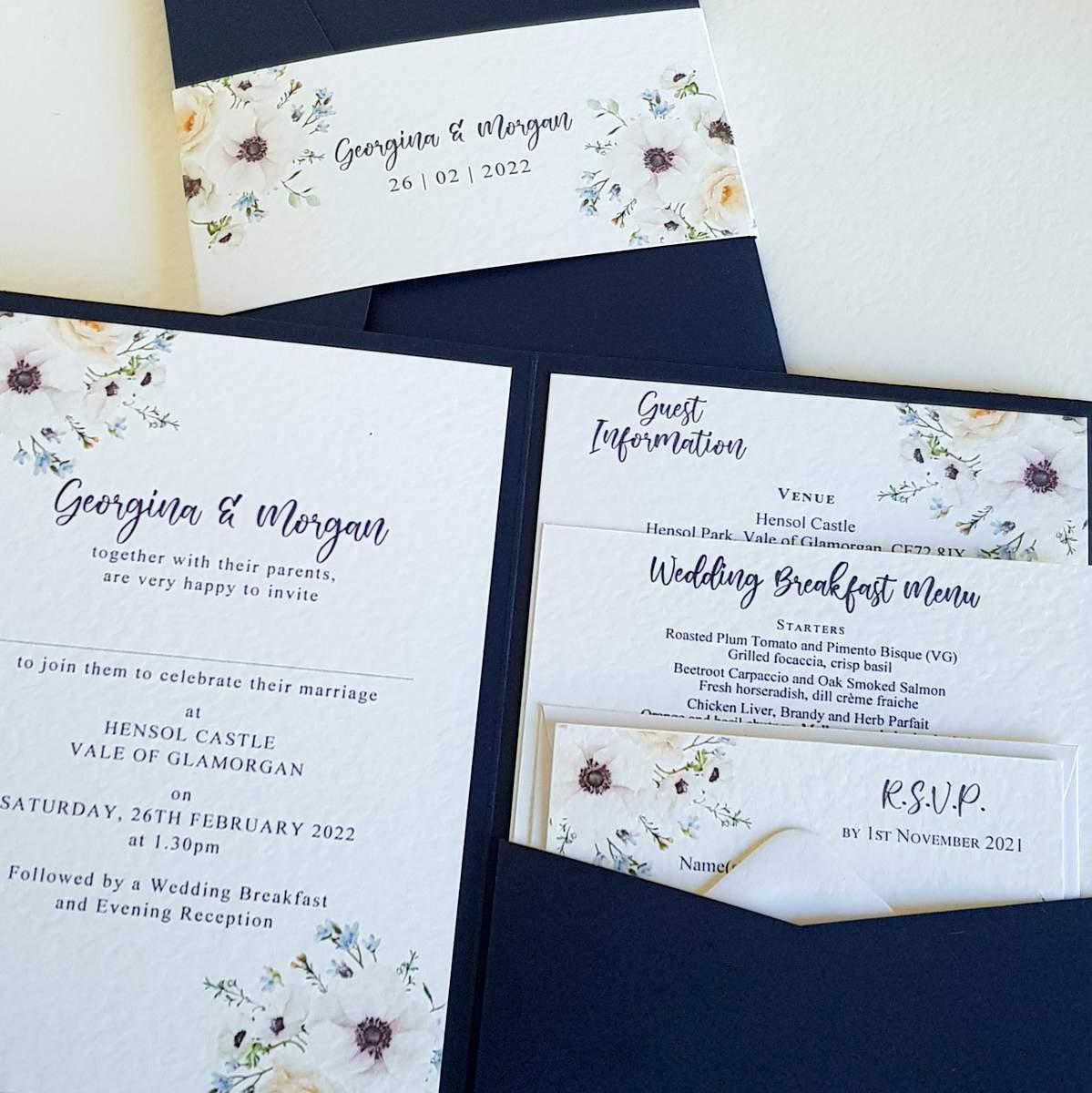 wedding invitations, an open pocketfold showing the invitation panel and the information cards inside the pocket. The outer pocket is navy blue and the cards inside are white, printed with a floral anemone design