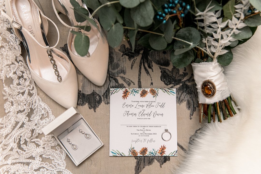 a winter wedding flat lay with invitation in a pine cone design, bridal shows, wedding bouquet and earrings in a box