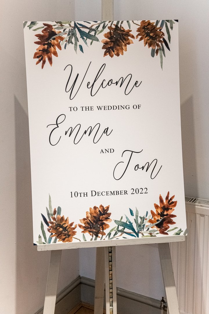 a sign with the words welcome to the wedding of emma and tom, greenery and pine cone motifs above and below the wording
