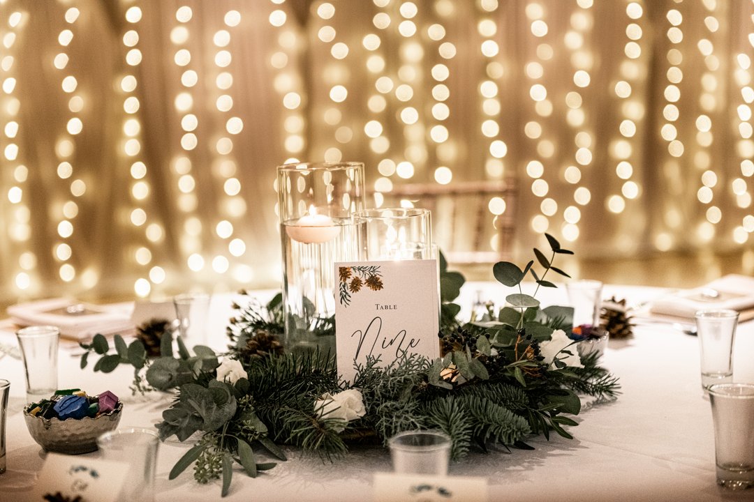 a wedding table setting with a printed table number showing a pine cone design, fairly lights and greenery