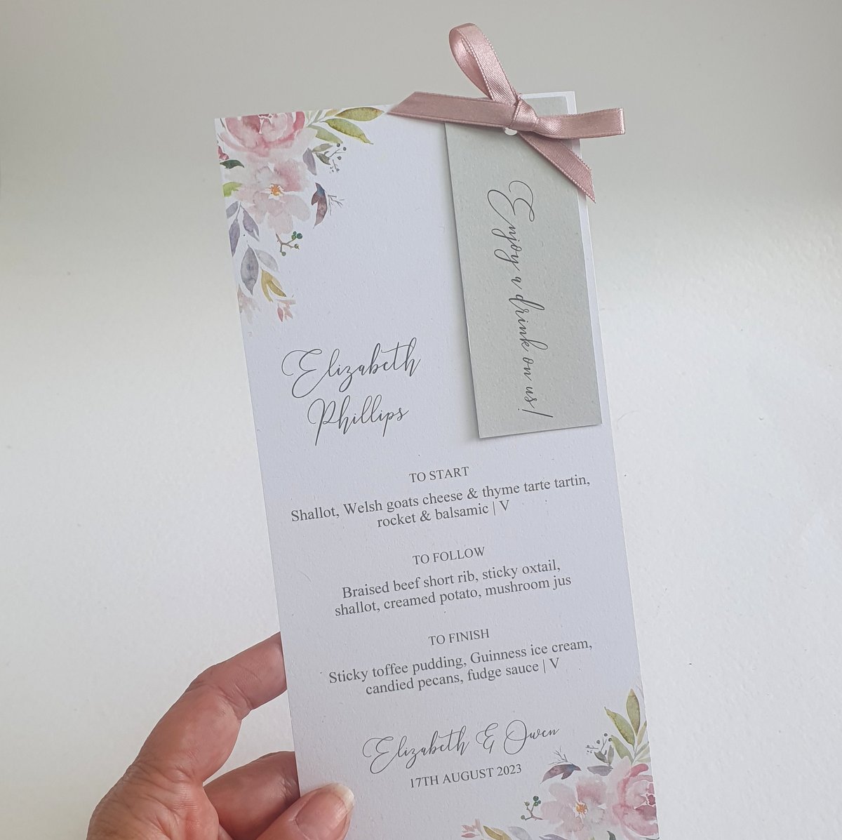 a personalised wedding menu with mauve floral accents, a silver grey name tag is attached with rose gold coloured satin ribbon in a bow