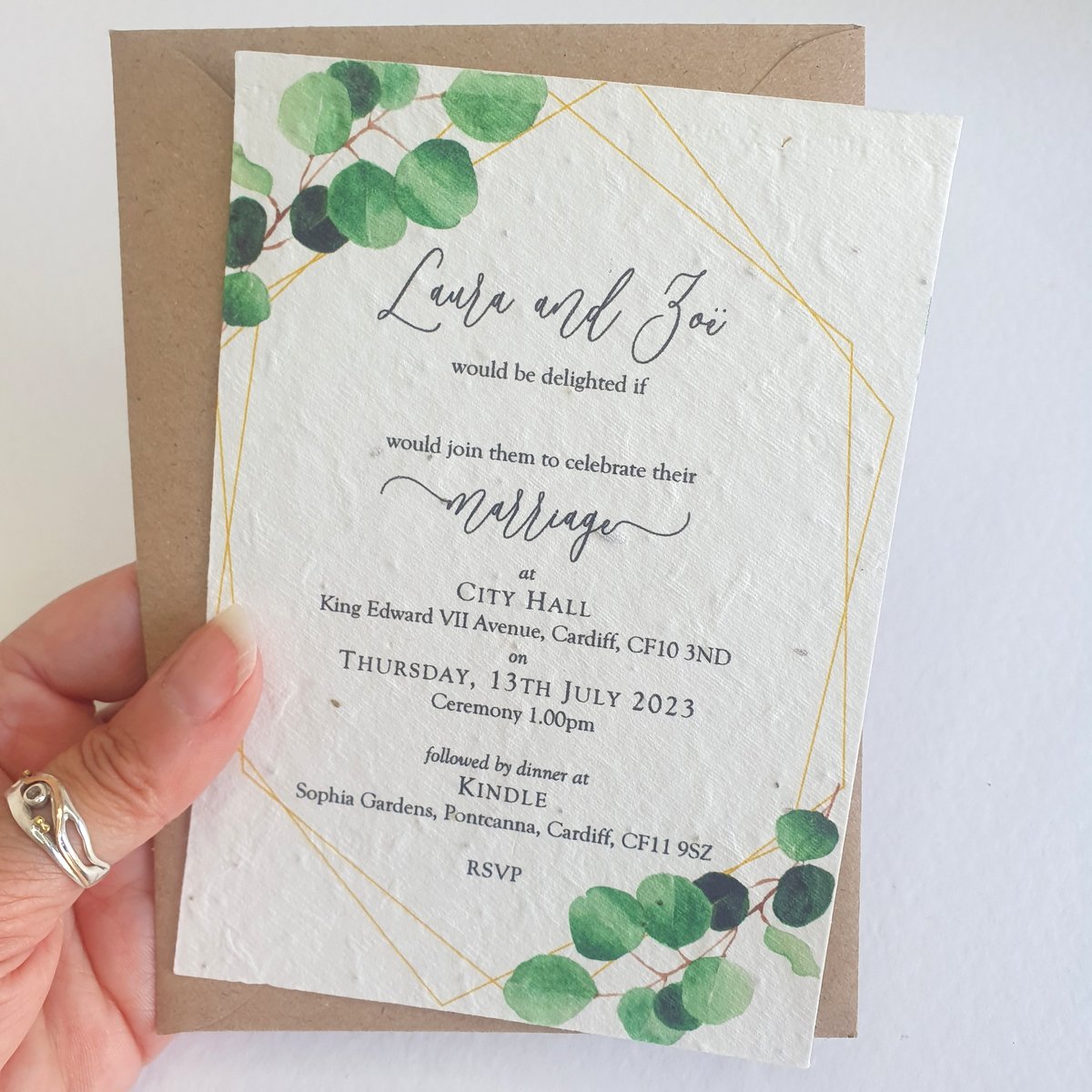 a wedding invitation with green eucalyptus leaves printed on the corners. The invitation is printed on seed paper and is shown with a recycled kraft brown envelope